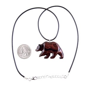 A dark reddish-brown wooden bear pendant necklace. The bear pendant is carved in a walking pose and hangs from a black cord necklace. A quarter is next to the pendant for size comparison.