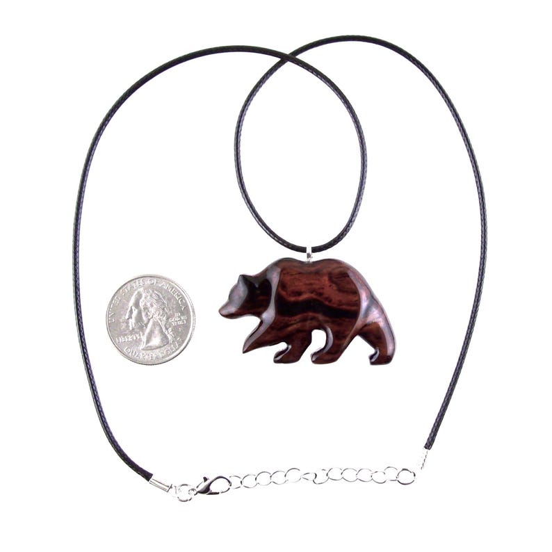 A dark reddish-brown wooden bear pendant necklace. The bear pendant is carved in a walking pose and hangs from a black cord necklace. A quarter is next to the pendant for size comparison.