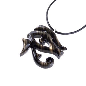 A wooden pendant necklace in black and brown, with a intricate design. The pendant is shaped like an Eye of Horus and hangs from a black cord.
