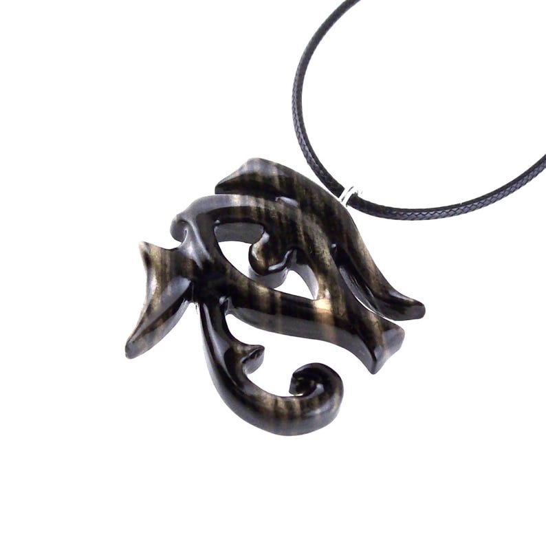 A wooden pendant necklace in black and brown, with a intricate design. The pendant is shaped like an Eye of Horus and hangs from a black cord.