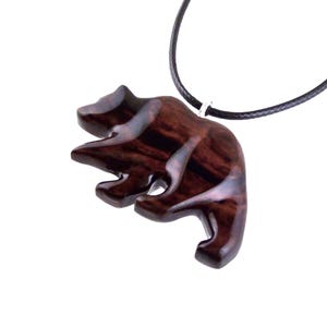 A wooden bear pendant with a dark reddish-brown finish and a black cord necklace. The bear is carved in a stylized form, with visible wood grain. The pendant is suspended from a black cord.