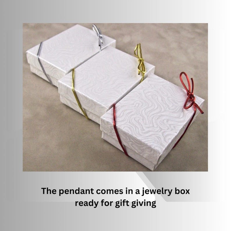 Three white jewelry boxes with different colored ribbon accents; silver, gold, and red.  The boxes have a subtle textured pattern and are ready for gift-giving. The text on the image reads: 'The pendant comes in a jewelry box ready for gift giving'.