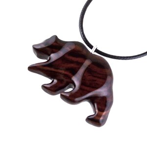 A wooden bear pendant with a dark reddish-brown finish and a black cord necklace. The bear is carved in a stylized form, with visible wood grain. The pendant is suspended from a black cord.