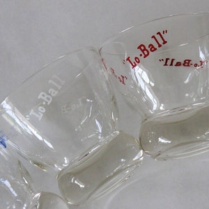 May include: Four clear glass drinking glasses with a rounded bottom and a wide mouth. Each glass has the words "Lo-Ball" printed on the side in white lettering with a red outline.