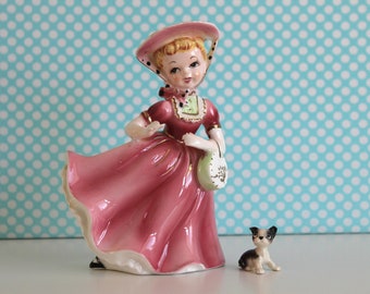 May include: A porcelain figurine of a girl in a pink dress and a hat, holding a basket. She is standing next to a small porcelain figurine of a black and white dog.