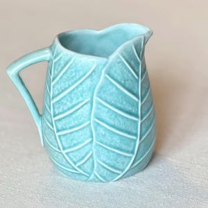 May include: A light blue ceramic pitcher with a leaf design. The pitcher has a handle and a spout. The leaf pattern is raised, creating a textured surface. The pitcher is approximately 6 inches tall.