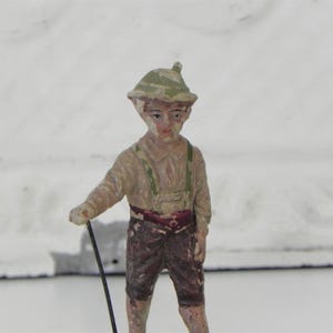 May include: A small, painted figurine of a boy wearing a green and brown hat, a brown shirt, and brown pants. He is holding a stick in his right hand.