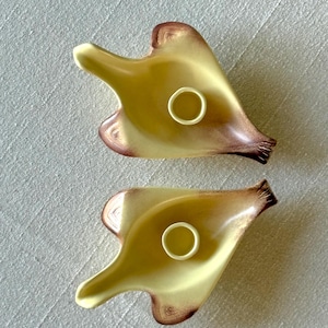 May include: Two yellow and brown ceramic ashtrays with a unique, organic shape. Each ashtray features a circular indentation and a flared, petal-like design. The ashtrays have a glossy finish and a vintage aesthetic.