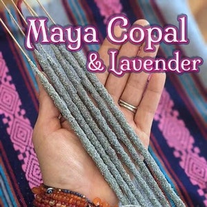 May include: A hand holding a bundle of gray incense sticks with the text "Maya Copal & Lavender" printed on a pink and blue background.