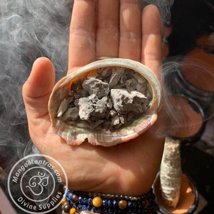 May include: A hand holding a seashell with grey stones inside. Smoke is rising from the stones. The hand is wearing several bracelets, including one with a blue evil eye charm.