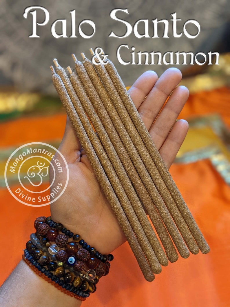 May include: A hand holds a bundle of eight brown Palo Santo & Cinnamon incense sticks. The sticks are about 10 inches long and have a rough texture. The background is a bright orange fabric.