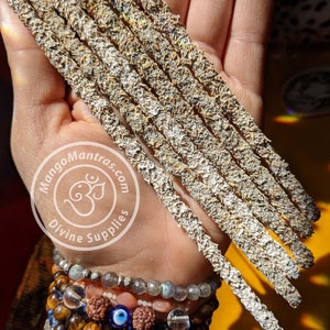 May include: A hand holding a bundle of eight incense sticks. The sticks are a light brown color and have a rough texture. The hand is wearing several bracelets, including a blue and white beaded bracelet, a brown and gold beaded bracelet, and a silver bracelet with a blue eye charm. The background is a yellow and orange color.