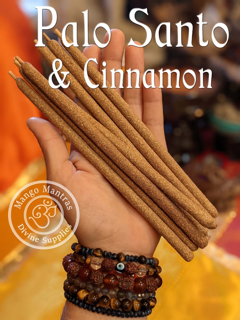 May include: A hand holds a bundle of brown Palo Santo & Cinnamon incense sticks. The sticks are about 10 inches long and have a rough texture. The hand is wearing several bracelets, including one with an evil eye charm. The background is blurred and out of focus.
