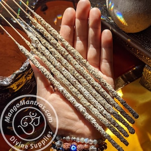 May include: A hand holding a bundle of eight incense sticks. The sticks are gray and white with a rough texture. The sticks are about 10 inches long. The hand is wearing several bracelets, including one with a blue evil eye charm. The image also includes a logo for Mango Mantras Divine Supplies.