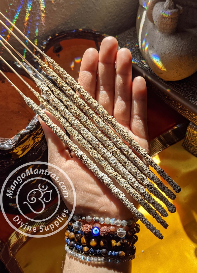 May include: A hand holding a bundle of eight incense sticks. The sticks are gray and white with a rough texture. The sticks are about 10 inches long. The hand is wearing several bracelets, including one with a blue evil eye charm. The image also includes a logo for Mango Mantras Divine Supplies.