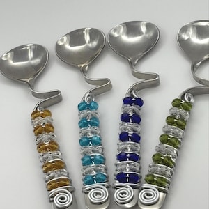 May include: Four silver spoons with beaded handles. The spoons have a unique curved design and are decorated with clear glass beads and colored glass beads in shades of blue, green, and gold.