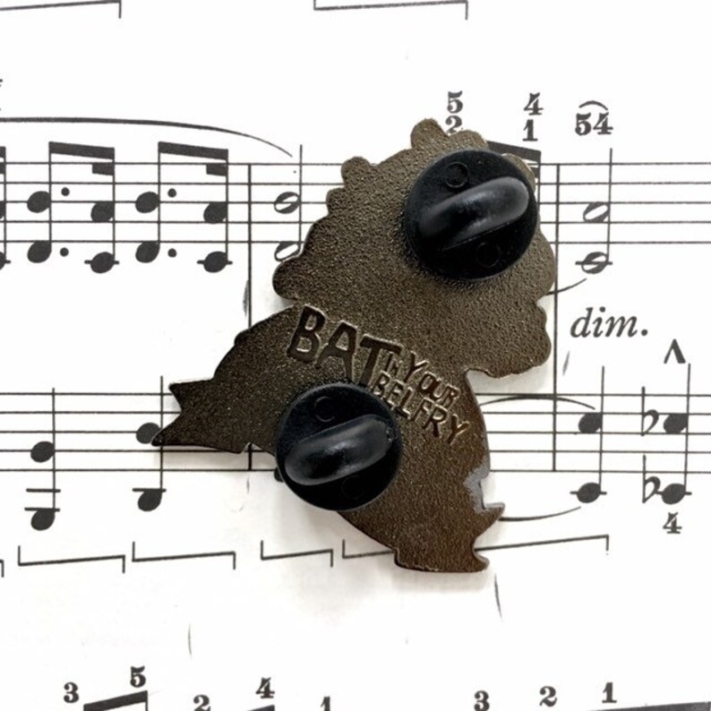 May include: A bronze-coloured enamel pin shaped like a bat, with two black pin backs. The text "BAT YOUR BELFRY" is visible on the pin. The pin is set against a backdrop of sheet music.