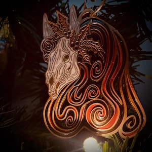 May include: A black and red metal ornament shaped like a horse's head with a skull-like face. The horse's mane is made of swirling lines and the horse is wearing a wreath of leaves and berries.