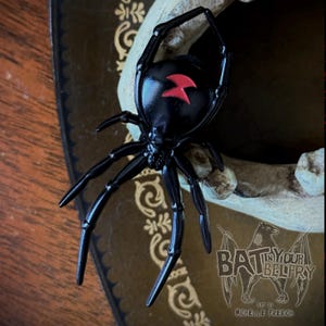 Op de afbeelding: Zwarte spinnenfiguur met een rode zandlopermarkering op de rug. De spin heeft acht poten en is geplaatst op een decoratief oppervlak. De afbeelding bevat ook de tekst "BAT IN YOUR BELFRY" en "ART BY MICHELLE PREBICH".