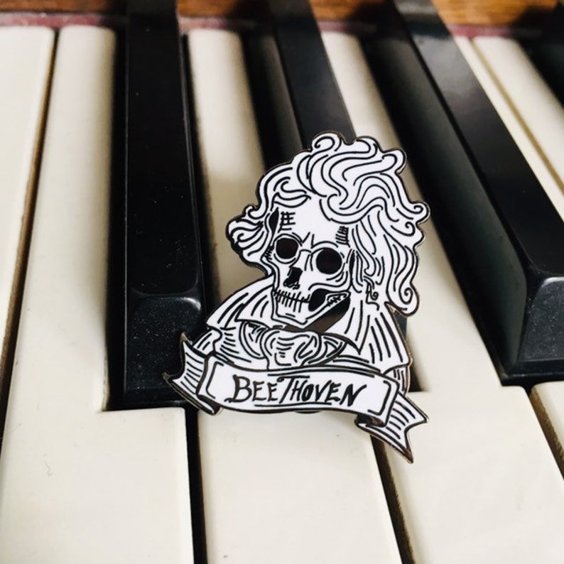 May include: A black and white enamel pin depicting a skull with Beethoven's hair and the word "BEE/THOVEN" on a banner. The pin is placed on a piano keyboard.