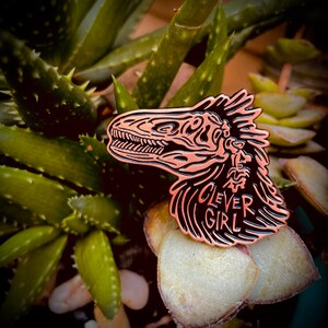 Peut inclure: Une &eacute;pingle en cuivre repr&eacute;sentant un cr&acirc;ne de dinosaure avec le texte "Clever Girl" en dessous. L'&eacute;pingle est pos&eacute;e sur une plante succulente verte.