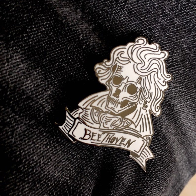 May include: A silver enamel pin depicting a skeleton with curly hair and a jacket, holding a banner with the word "Beethoven". The pin is set against a dark denim background.