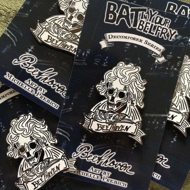 May include: Black and white enamel pins with a skeletal Beethoven design, labelled "BeeThoven" on a banner. The pins are part of the "Decomposer Series" by Michelle Prebich. The background is a dark blue with a subtle pattern.
