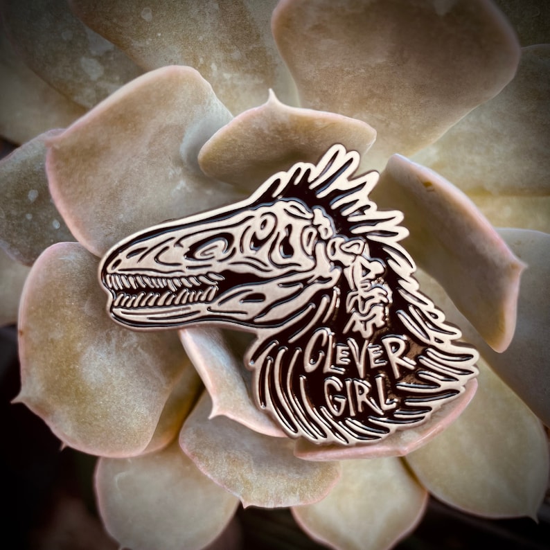 Peut inclure: Une &eacute;pingle en &eacute;mail noir et argent repr&eacute;sentant un cr&acirc;ne de dinosaure stylis&eacute; avec le texte "Clever Girl" en dessous.