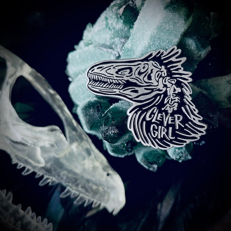 Peut inclure: &Eacute;pinglette en &eacute;mail noir et blanc repr&eacute;sentant une t&ecirc;te de dinosaure stylis&eacute;e avec le texte "Clever Girl" en dessous. L'&eacute;pingle est sur un fond vert et bleu.