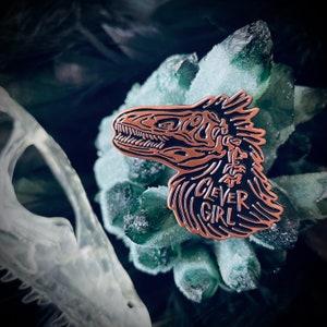 Peut inclure: Une &eacute;pingle en &eacute;mail de couleur cuivre repr&eacute;sentant une illustration d&eacute;taill&eacute;e d'un dinosaure avec le texte "Clever Girl" en dessous.