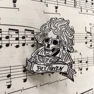 May include: A black and white enamel pin depicting a skull in the style of Beethoven, with the word "Beethoven" on a banner. The pin is set against a backdrop of musical notation.