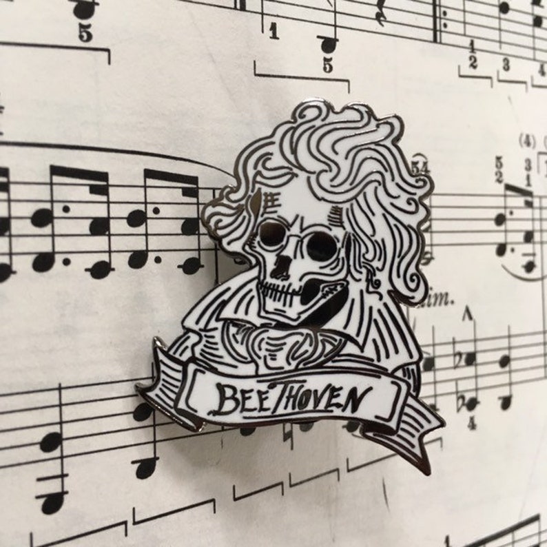 May include: A black and white enamel pin depicting a skull in the style of Beethoven, with the word "Beethoven" on a banner. The pin is set against a backdrop of musical notation.