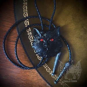 May include: A black bolo tie featuring a cat's head with red eyes. The cat pendant is detailed and has a textured surface. The bolo tie has a braided black cord and a decorative metal slide.