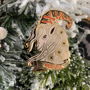 May include: A gold-colored enamel pin featuring a white goat with red and gold horns. The goat is surrounded by a white and gold swirl design. The pin has a banner at the bottom that reads "God Jul" in red and gold lettering.
