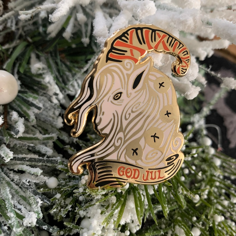 May include: A gold-colored enamel pin featuring a white goat with red and gold horns. The goat is surrounded by a white and gold swirl design. The pin has a banner at the bottom that reads "God Jul" in red and gold lettering.