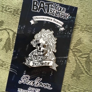 May include: A black and white enamel pin depicting a skull with curly hair, a banner reading "Beehoven," and the text "Bat in Your Belfry." The pin is on a dark blue card with the artist's name.