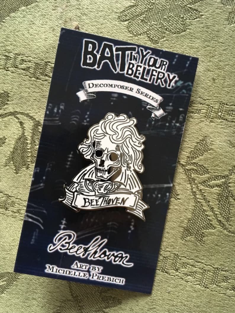 May include: A black and white enamel pin depicting a skull with curly hair, a banner reading "Beehoven," and the text "Bat in Your Belfry." The pin is on a dark blue card with the artist's name.