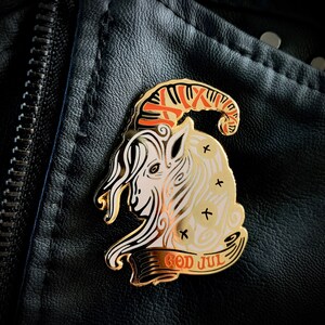 May include: Gold-colored pin with a white goat head with red and orange horns. The goat is surrounded by a banner that reads "God Jul" in gold lettering.