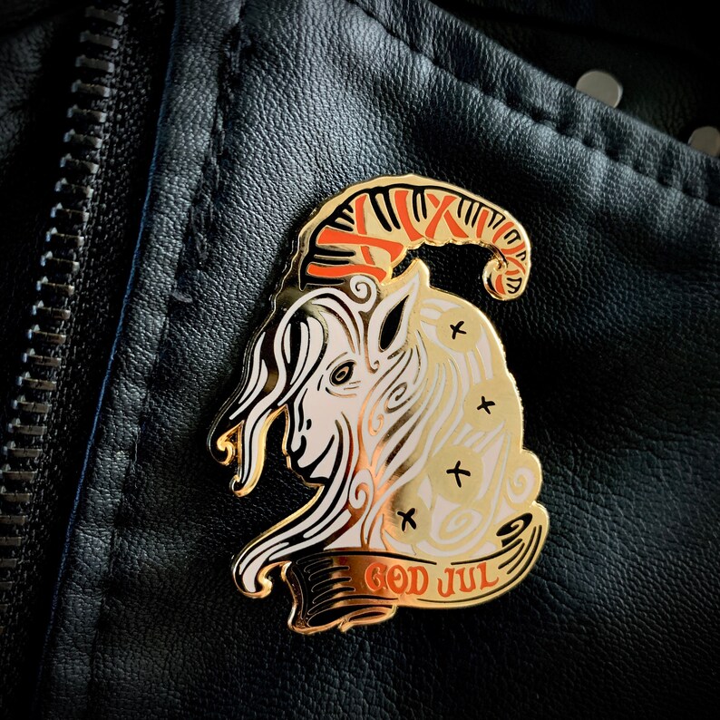 May include: Gold-colored pin with a white goat head with red and orange horns. The goat is surrounded by a banner that reads "God Jul" in gold lettering.