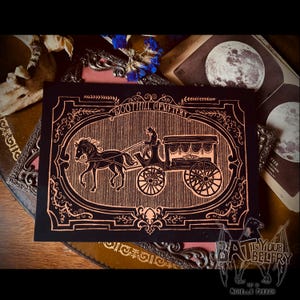 May include: A rectangular black print featuring copper-colored line art of a horse-drawn hearse. The central illustration is enclosed in an ornate frame with the text BOOTHILL CEMETERY arched above. The design depicts a driver, a horse, and a carriage.