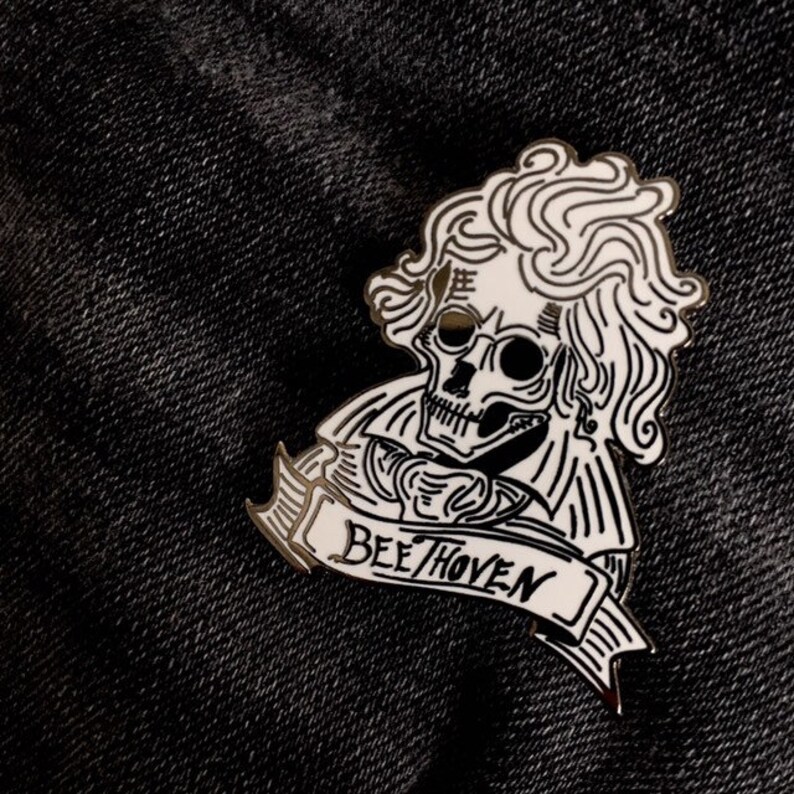 May include: A monochrome enamel pin depicting a skull in the style of Beethoven, with the name "Beethoven" on a banner. The pin is set against a dark denim background.