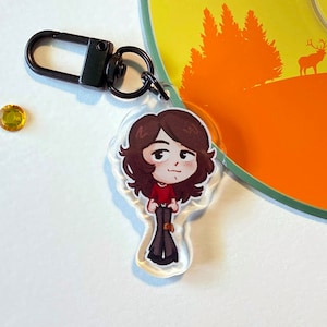 Puede incluir: Un llavero negro con un dije de acrílico transparente que presenta una ilustración de dibujos animados de una persona con cabello largo castaño que lleva una camisa roja y pantalones negros.