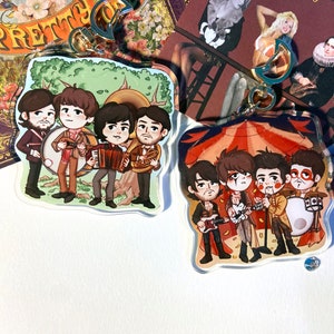 May include: Two colorful acrylic keychains featuring cartoon illustrations of a band. The first keychain shows four musicians playing instruments in a forest setting. The second keychain shows four musicians playing instruments in a circus tent.
