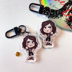Peut inclure: Deux porte-clés noirs avec des breloques en acrylique transparent représentant une illustration de personnage de dessin animé avec des cheveux foncés, portant une chemise noire, un short noir et des chaussures noires. La personne fait un doigt d'honneur avec les deux mains.