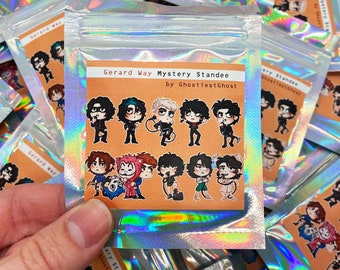 Puede incluir: Una bolsa holográfica transparente que contiene un conjunto de pegatinas. Las pegatinas presentan ilustraciones de dibujos animados de una persona en varias poses. El texto "Gerard Way Mystery Standee" y "by GhostiestGhost" está impreso en una etiqueta.