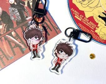 May include: Two clear acrylic keychains featuring cartoon illustrations of a person with brown hair, wearing a red shirt, black pants, and a white collar. The keychain is attached to a black metal ring and a black metal clasp. The background is a white surface.