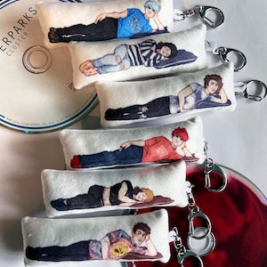 May include: Six white fabric keychains with different illustrations of people lying down. Each keychain has a silver metal ring and a silver metal hook.