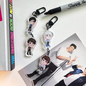 May include: Four clear acrylic keychains with cartoon illustrations of a person, each attached to a black metal clasp. A black Sharpie marker and a photograph of a person are also in the image. The keychains are on a white surface.