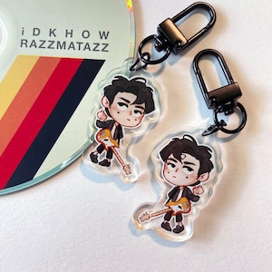 Puede incluir: Dos llaveros de acrílico transparente con ilustraciones de dibujos animados de una persona con cabello negro, vistiendo una chaqueta negra y sosteniendo una guitarra amarilla. Los llaveros están unidos a llaveros de metal negro.
