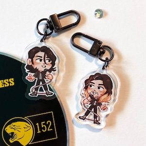 Puede incluir: Dos llaveros de acrílico transparente con anillas de metal negro. Cada llavero presenta una ilustración de dibujos animados de una persona sosteniendo un micrófono. Una persona tiene el pelo negro y la otra tiene el pelo castaño con reflejos rubios.